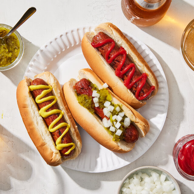 A paper plate with three hot dogs with toppings is surrounded by those condiments and glasses of beer.