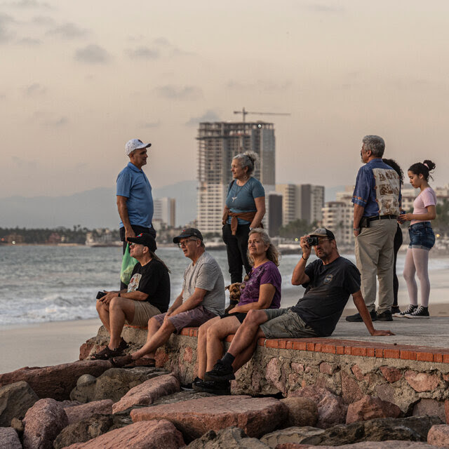 A group of people sitting on a wall next to the beach with construction in the background.