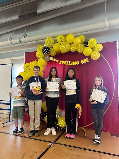 Five children stand in front of a "Spelling Bee" banner and balloon decorations, holding certificates and trophies. They are on a stage inside a room with bright lighting and large windows.