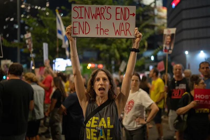 A woman holds up a sign that says Sinwar's End --> End the War amid a crowd of protesters on a city street at night.