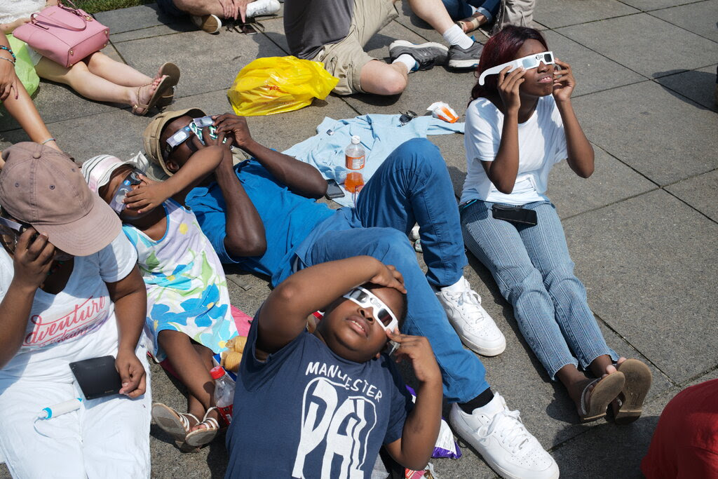 People lie on the ground in a large gathering to observe the solar eclipse, all of them holding protective glasses over their eyes.