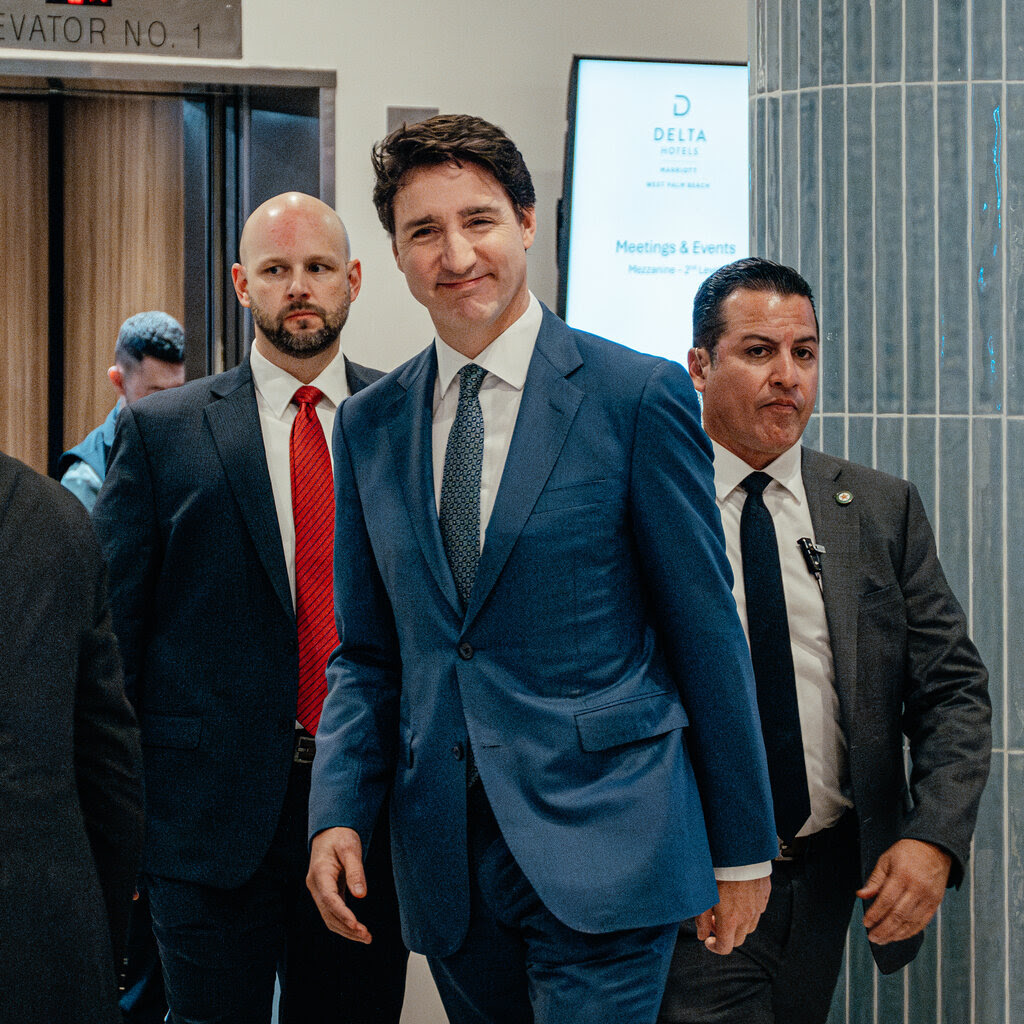 Justin Trudeau, wearing a blue suit and smiling, walks through a hotel lobby, flanked by men who look like security guards.