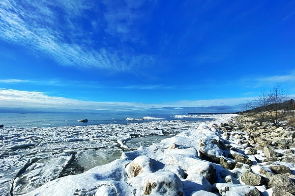 snow-covered rocks on shore and calm, gray-blue water stretch out beneath a brilliant blue winter sky framed by a few white, wispy clouds