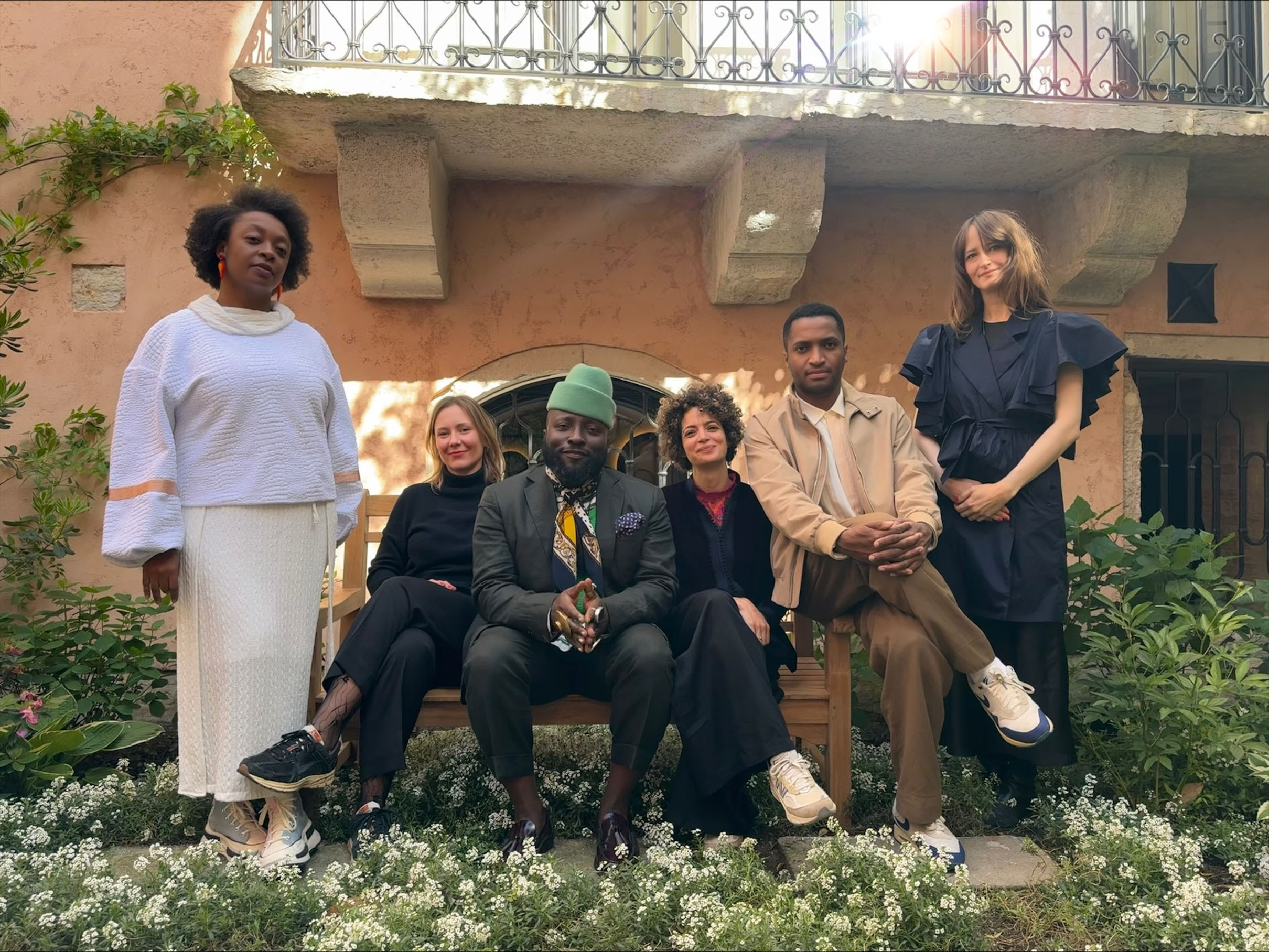 A foto mostra seis pessoas lado a lado num jardim com uma varanda atrás e um raio de luz passando por elas – algumas de pé e algumas sentadas –, ao lado esquerdo está uma mulher negra de cabelo afro vestida de branco de pé, uma mulher branca e loira sentada sorrindo vestida de preto, um homem negro de terno, gorro verde e lenço no pescoço sorrindo e juntando as mãos, uma mulher de origem árabe e cabelo encaracolado vestida de preto sorrindo, um homem negro de cabelo curto vestido de bege cruzando as mãos sobre as pernas, e uma mulher branca de vestido preto longo sorrindo de pé.