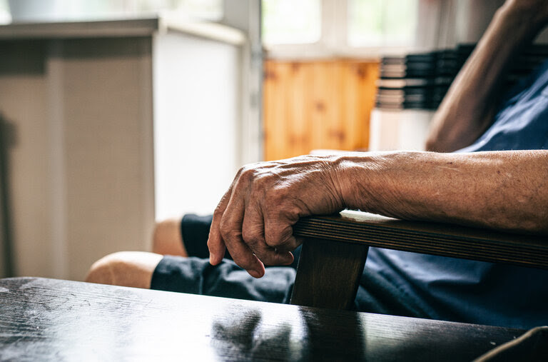 Cropped wrinkled senior man hand holding armrest of the chair in bright room.