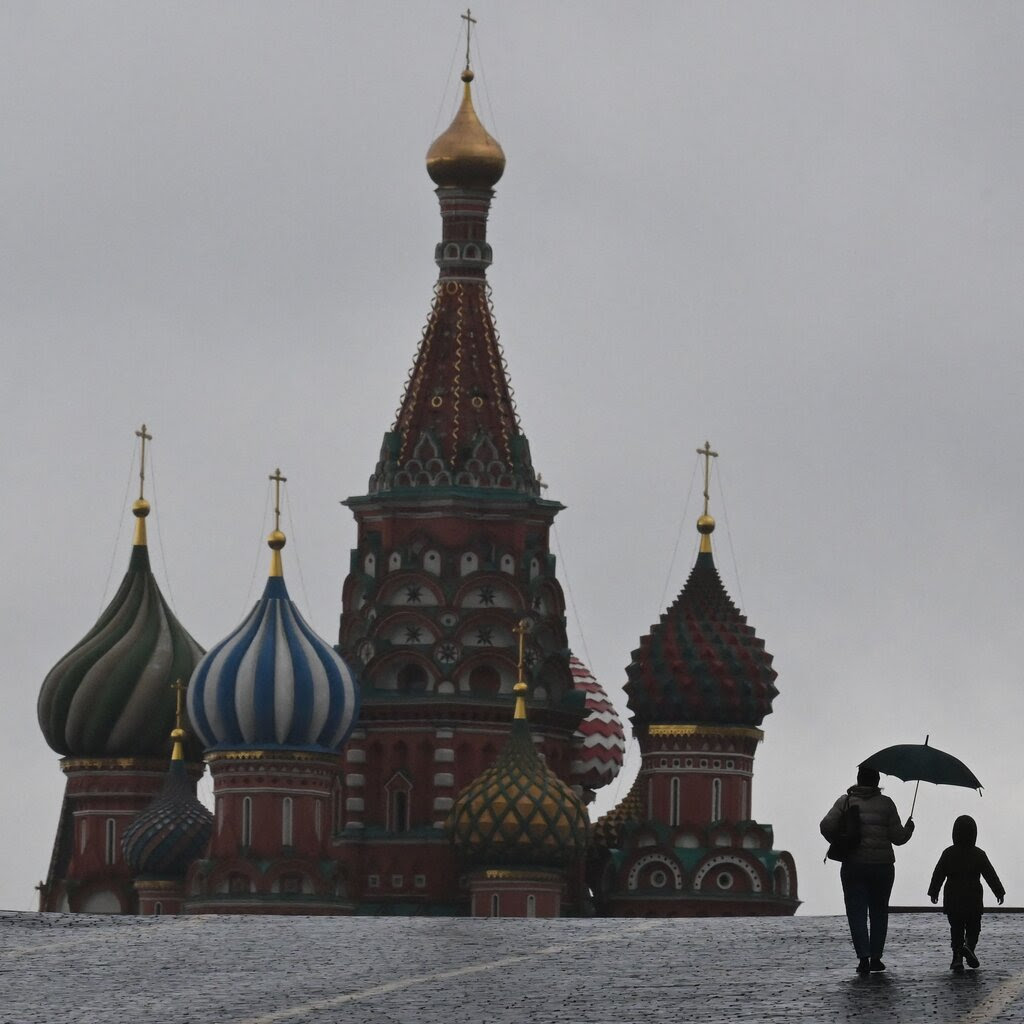 An adult with an umbrella walking with a child is silhouetted against a gray sky on Red Square, with the domes of St. Basil’s Cathedral on the left. 