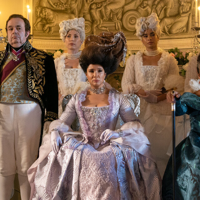 Six women and one man, all dressed in elaborate Regency period dress stand in two lines, many wearing white wigs. 