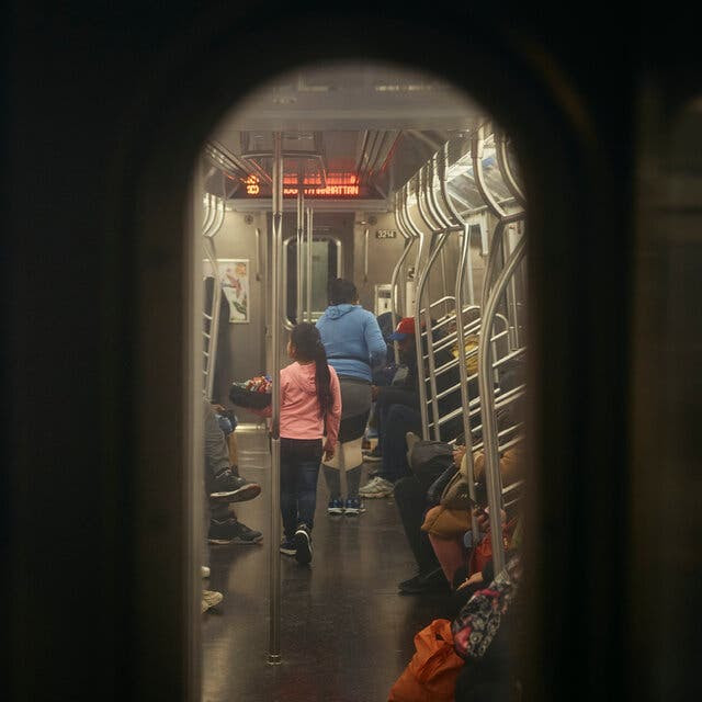 A young girl in a pink hooded sweatshirt is seen through an oval subway car window from the back. She carries a box of candy in her arms. Ahead of her is a woman in a blue hooded sweatshirt.