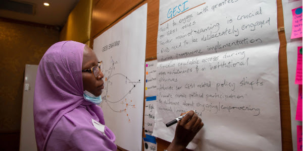 A woman in a purple headscarf and glasses takes notes on a writing pad during a gender equity and social inclusion training.