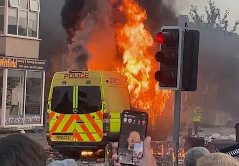 A photo of a police truck on fire, with big and high flames surrounded by trash and destruction caused by a riot in the UK, a crowd is watching.