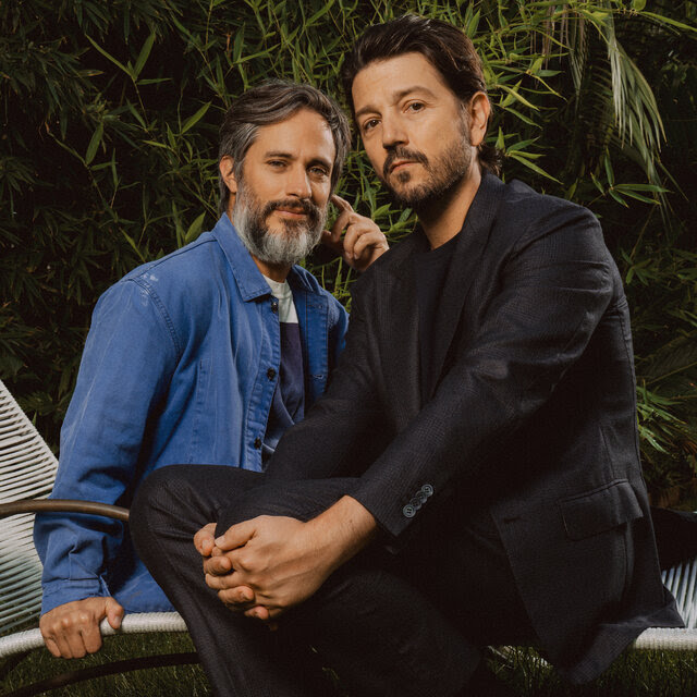 Two dark-haired men in an outdoor garden, one wearing a blue workshirt and the tother a suit with a black T-shirt, sit and pose for the camera. 