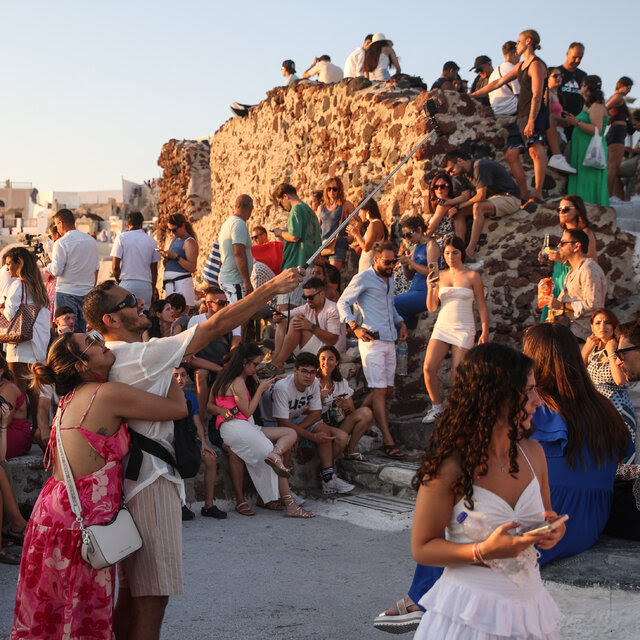 A large crowd of people in holiday clothing is gathered around a crumbling stone wall in the early evening, before sunset. In the foreground a man in shorts and sunglasses holds a selfie stick in the air as he takes a photo of himself and a young woman in a colorful sundress who is hugging him from behind. In the distance are whitewashed buildings. 