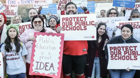Adults and children stand outside at a rally in support of public education.