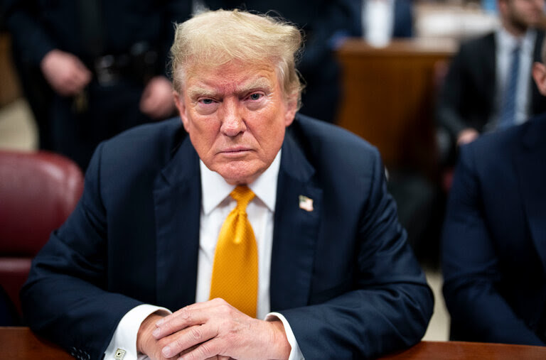 Donald J. Trump, wearing a navy suit and a yellow tie, scowls as he sits at a defense table.