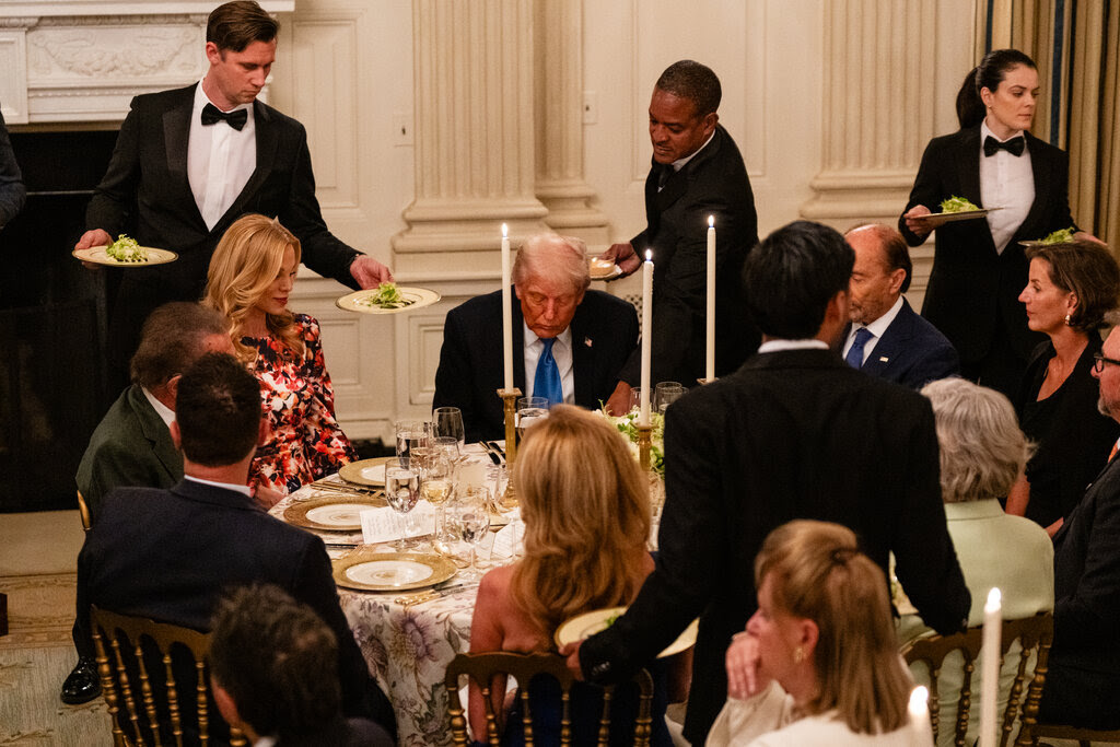 Waiters in black tuxedos serve plates of salad to President Trump and his guests seated at a round table for a candlelight dinner.