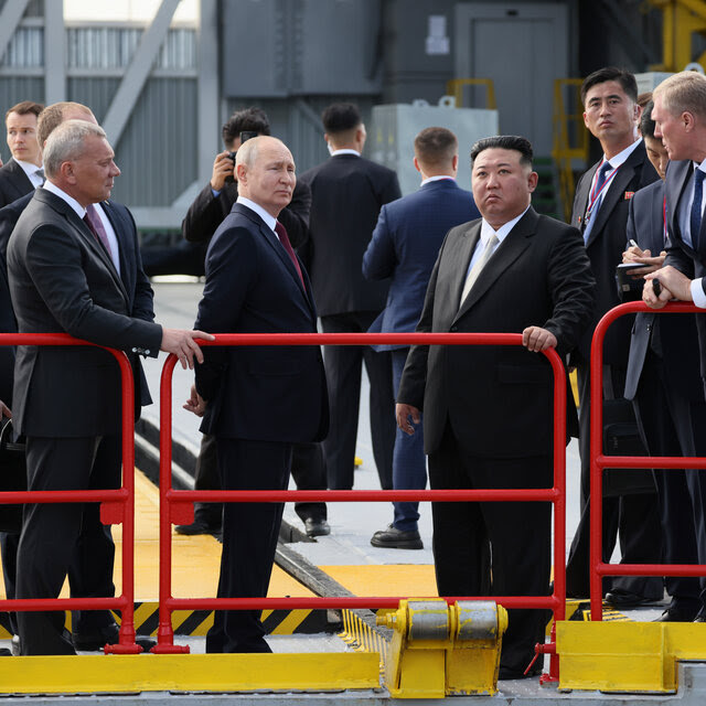 Vladimir Putin and Kim Jong-un, wearing suits, stand at a railing, with other men surrounding them.