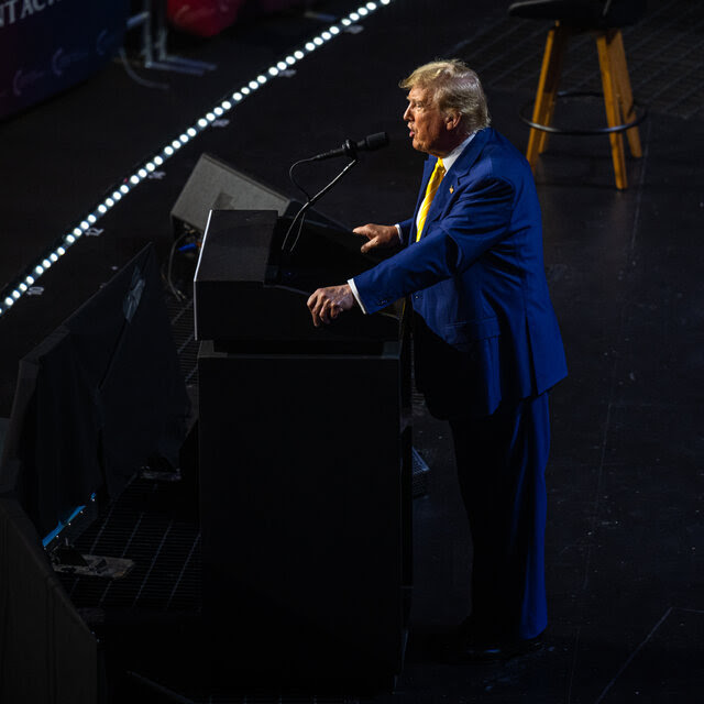 Former President Donald J. Trump, speaking on a stage behind a lectern and in a blue suit and yellow tie, facing left.
