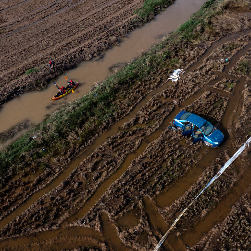 A blue car abandoned in a muddy, flooded field as rowers in a canoe pass by along a nearby water channel.