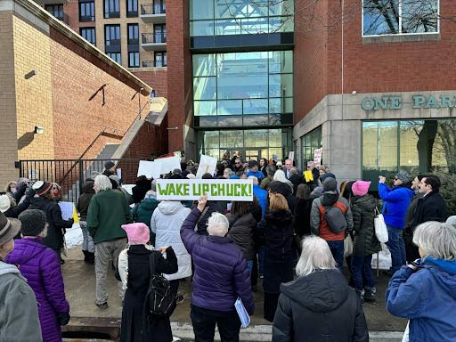 Activists outside an office building. One holds a sign that reads wake up, Chuck!