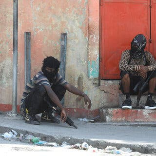 People with their faces covered, holding long guns while sitting on a step and the sidewalk.