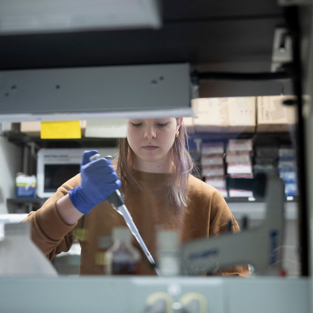 A researcher with a pipette and blue gloves in a lab space, seen through an opening between shelves.