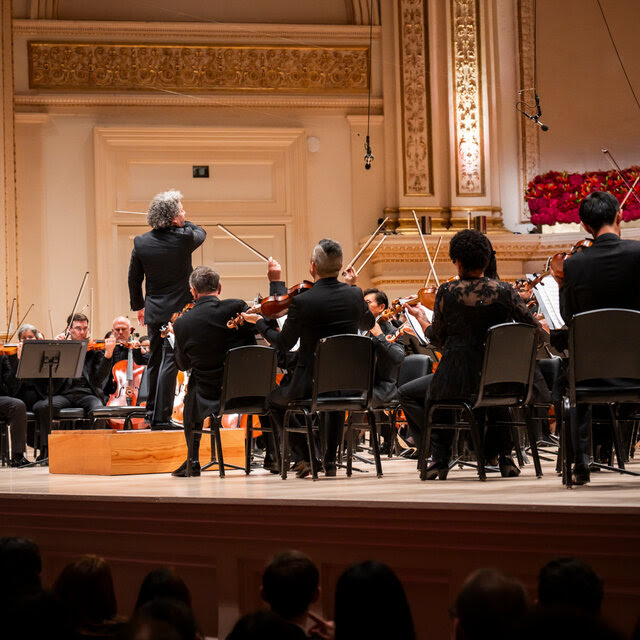 On the stage of Carnegie Hall, we see the conductor Gustavo Dudamel from the back, surrounded by string players. 