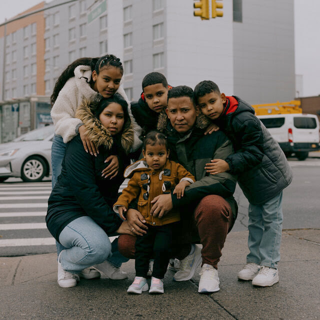 A family of six kneels on a New York City street corner wearing jackets and holding each other close