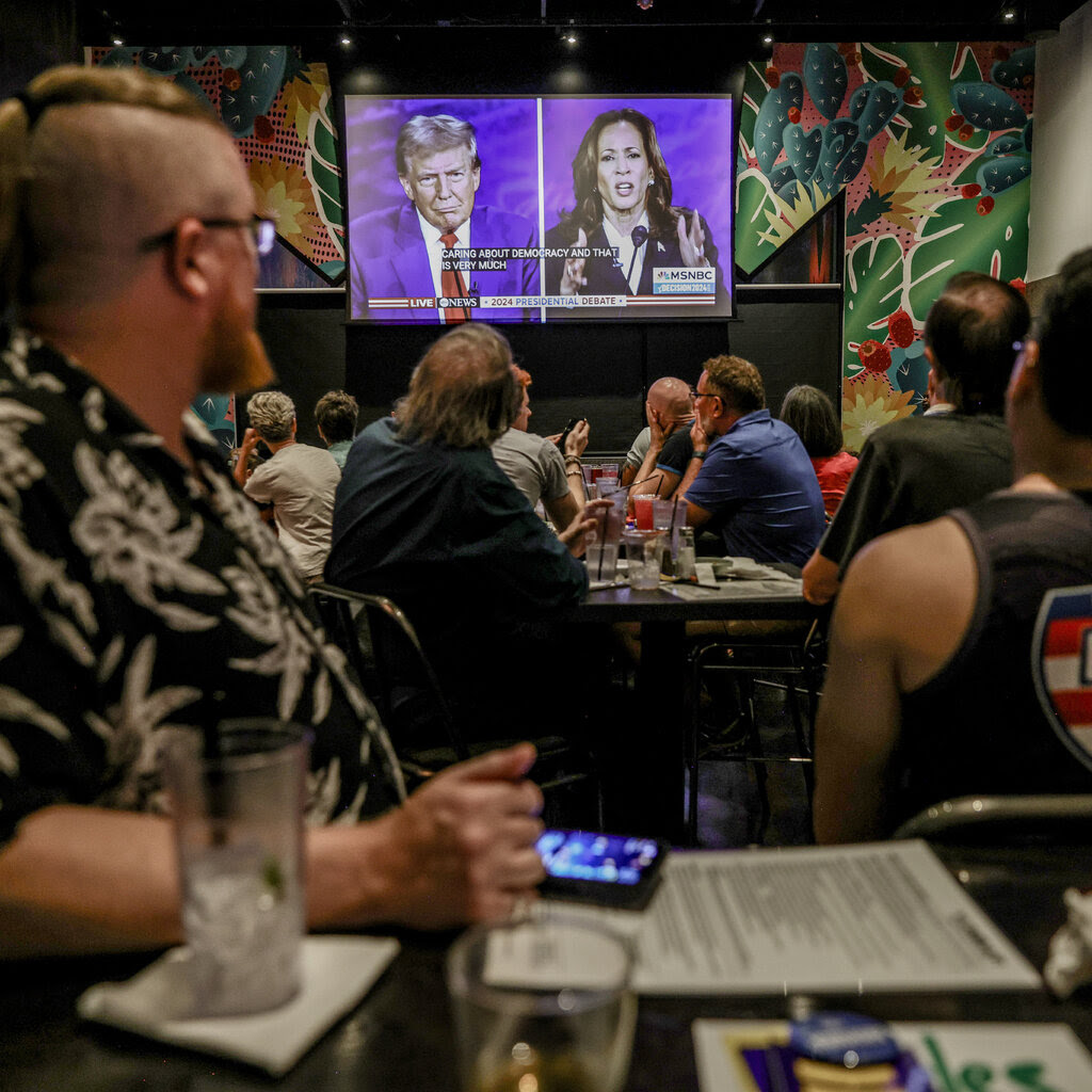 A seated crowd in a bar watching a split-screen from the debate on a wall-mounted television.