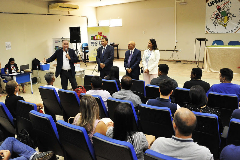 A imagem mostra um evento em um auditório, onde várias pessoas estão sentadas em cadeiras azuis, assistindo a uma apresentação. Quatro pessoas estão em pé na frente do auditório: três homens e uma mulher, todos vestidos formalmente. Um dos homens está falando ao microfone. No fundo, há um banner com o texto "REGIS PARA TODOS" e o logotipo da Universidade Federal do Maranhão (UFMA) na parede. À esquerda, uma mulher está sentada em uma mesa com um laptop.