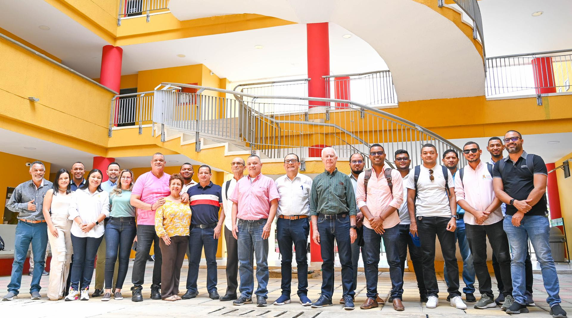 un grupo numeroso de personas posa juntas en lo que parece ser el patio de un edificio de varios niveles, con paredes de color amarillo brillante y columnas rojas. El grupo está compuesto por hombres y mujeres de diferentes edades, todos sonrientes, de pie y mirando hacia la cámara. Al fondo, se observa una estructura arquitectónica moderna con barandillas metálicas y escaleras en espiral que adornan el edificio.