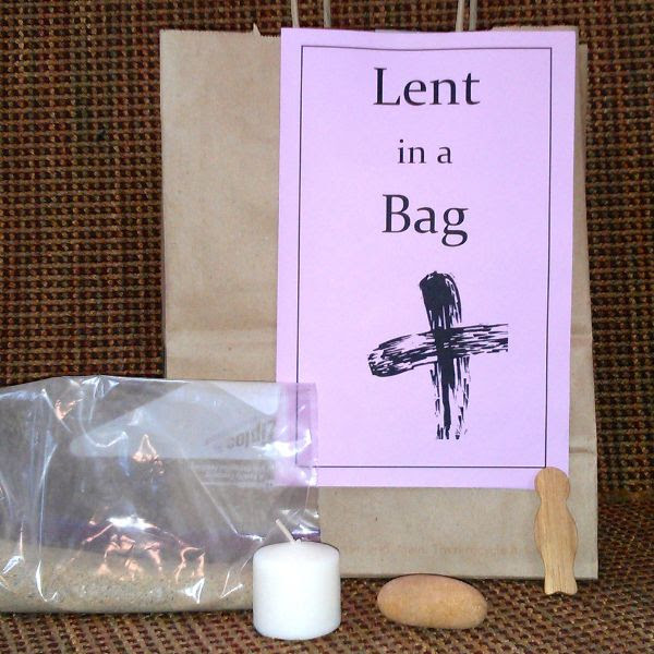 A piece of light purple paper that says "Lent in a Bag" with a cross symbol in black on a brown paper bag, a clear plastic bag with sand, an unlit white votive candle, a brown rock, and a brown wood figure on a dark brown background