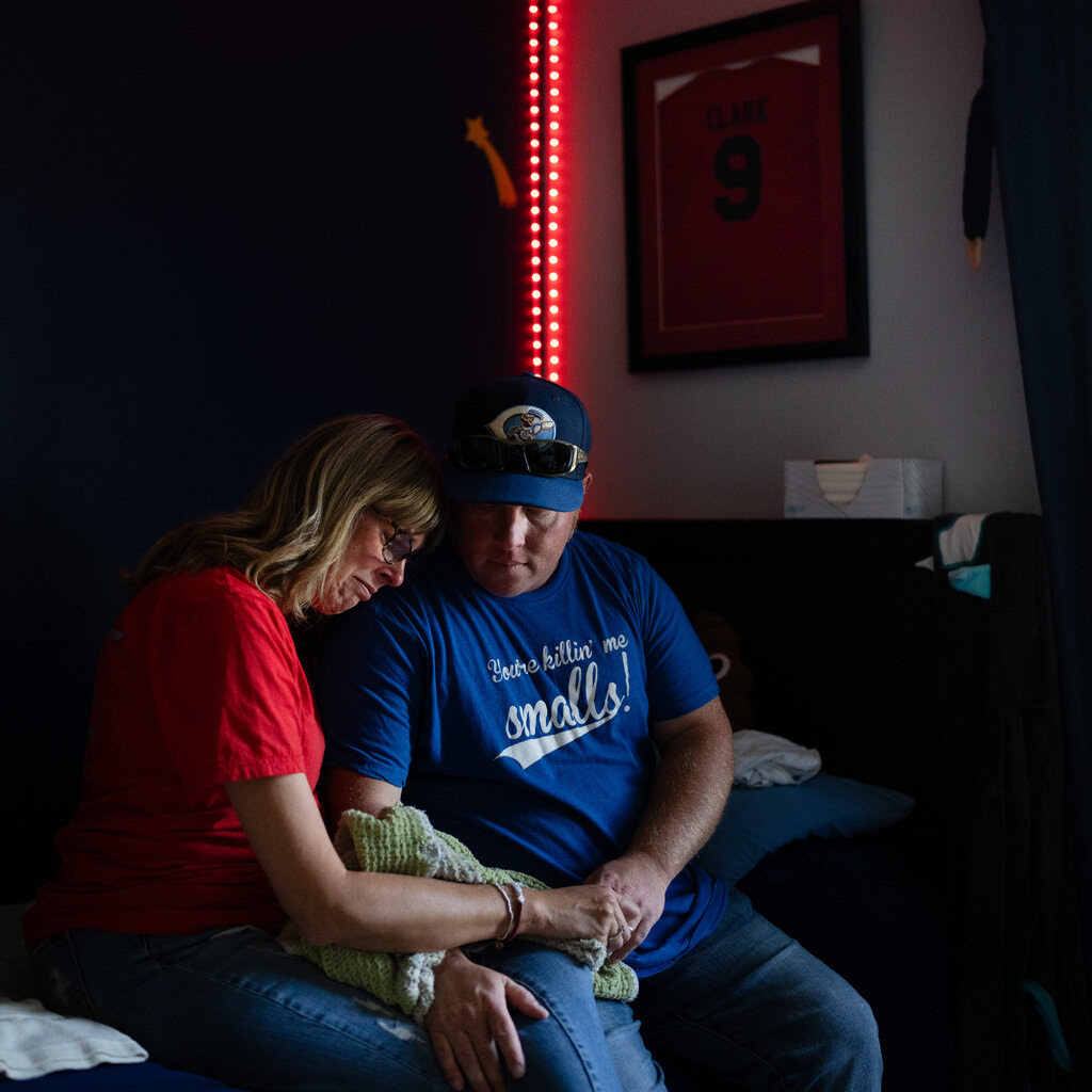 A man and woman sit, with their heads together, on the side of a bed. 