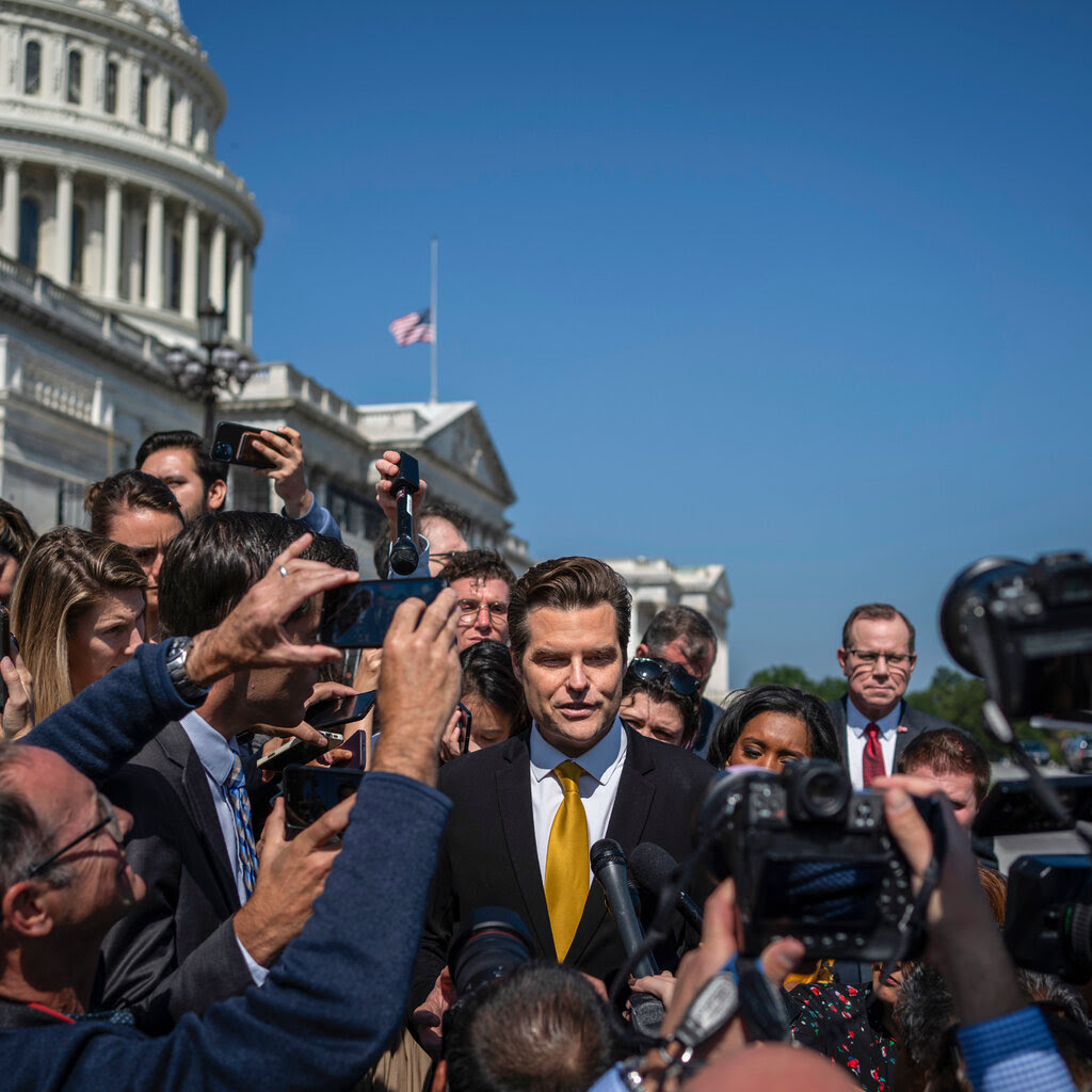 Gaetz, in a suit and yellow tie, walks through a crowd of reporters outside at the Capitol. 