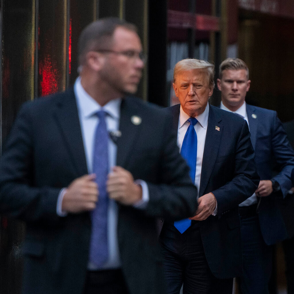 Donald Trump walks out of a doorway in a suit and a blue tie, with two security guards near him.