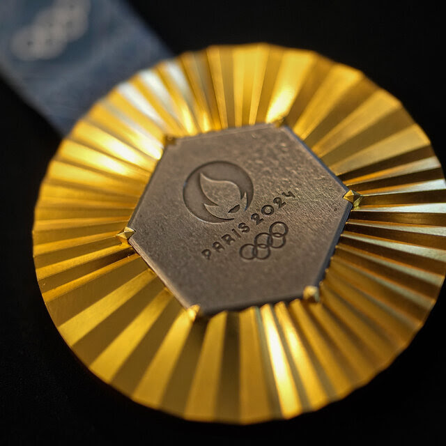 A gold medal with a piece of metal in the middle that says Paris 2024 above Olympic rings.