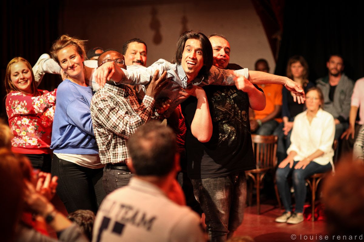 Six individuals, comprising men and women, carry a smiling, spreadeagled man during a performance. In the background, others watch on.