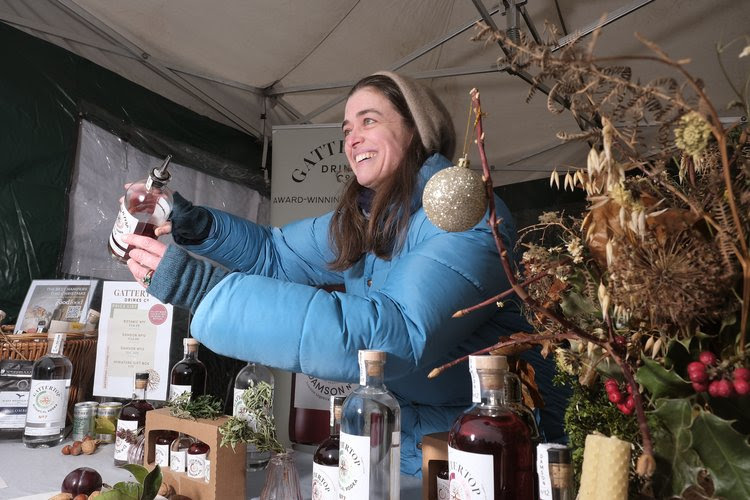 A smiling stall holder dressed in winter clothes pouring a sample of a drink for a customer.