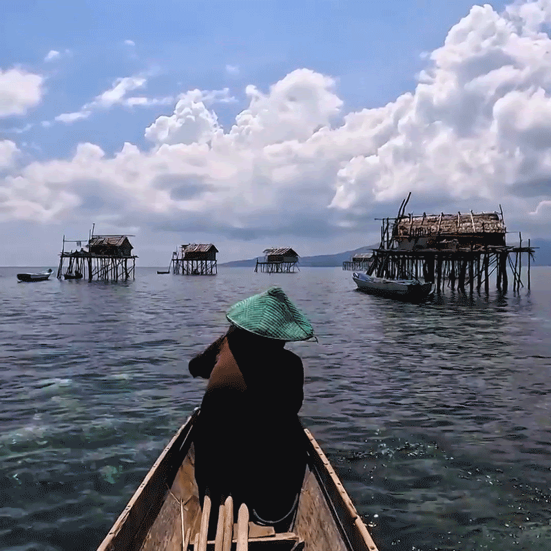 A person wearing a green conical hat in a wooden boat, floating on clear water, with elevated houses in the distance