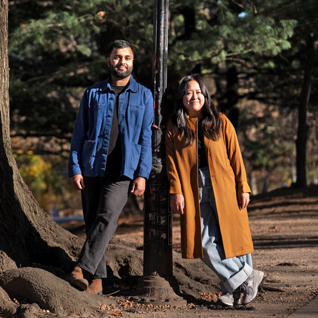 A man and woman stand outdoors, leaning on a lamp pole in a park. He wears a blue jacket and she wears a long brown coat.