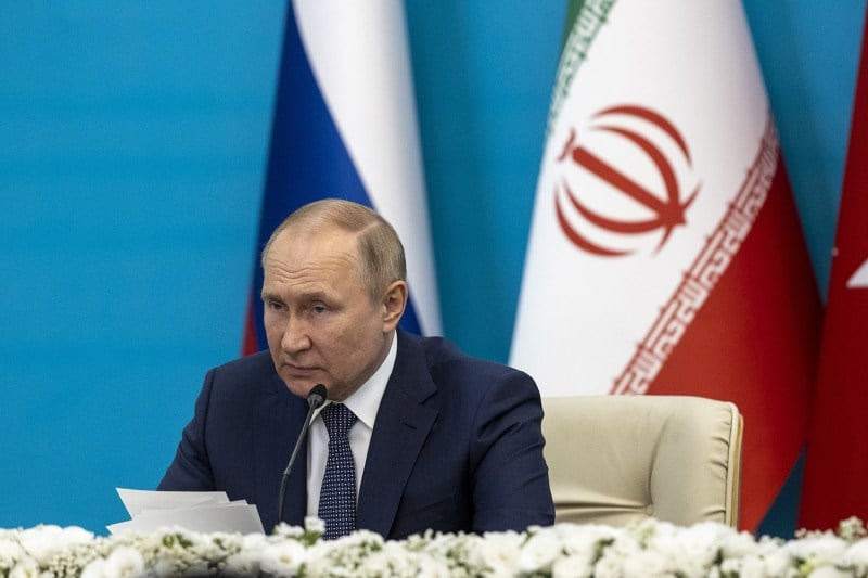 Russian President Vladimir Putin in a suit and tie sits in front of a microphone and table decorated with flowers. The Russian and Iranian flags are seen behind him.