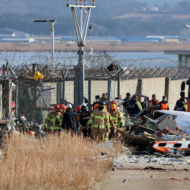 Next to a wall topped with razor wire, a group of people in firefighting uniforms stand near the wreckage of an airplane. 