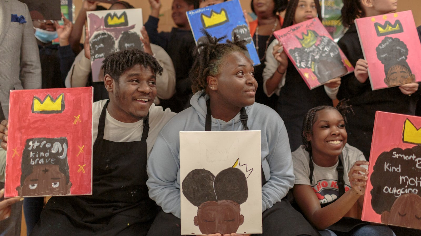 Several students wearing black smocks smile and hold up artwork featuring yellow crowns.