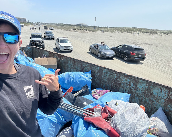 Volunteer perched atop a full dumpster with cars driving in and out of the beach approach at Ocean Shores 