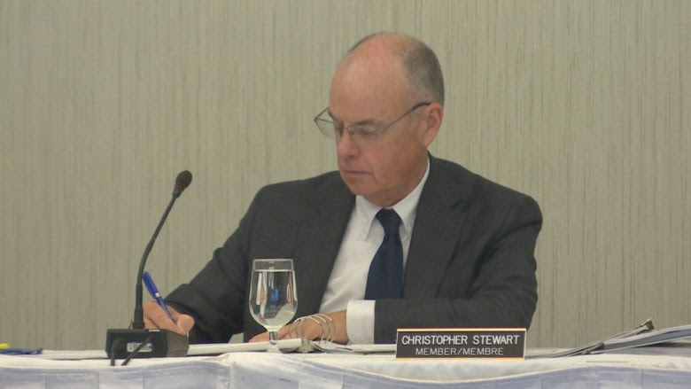 A bald man in a grey suit sits at a table behind a microphone and writes on a piece of paper.