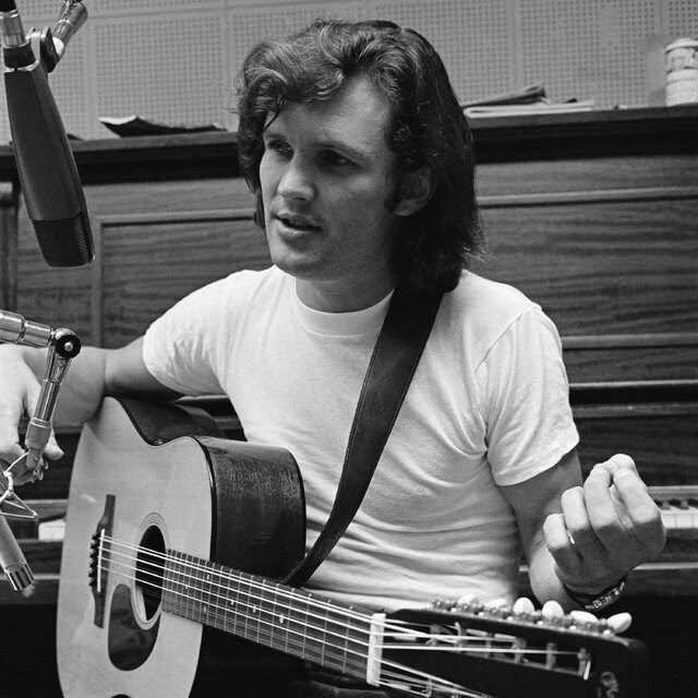 A black-and-white photo of Kris Kristofferson, in a white T-shirt, sitting on a piano bench faced away from the instrument, an acoustic guitar on his lap and a microphone poised in front of his face.