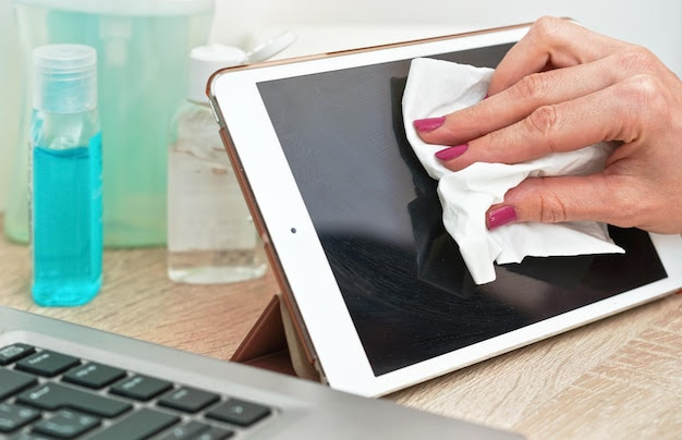 Woman cleaning tablet device with cotton paper tissue, wiping black screen, closeup detail. Blurred bottles with washing products background. Coronavirus covid-19 outbreak prevention