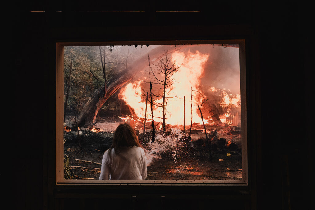 As flames get close to her property, a woman sprays water to keep the fire away, a view seen from inside her home.