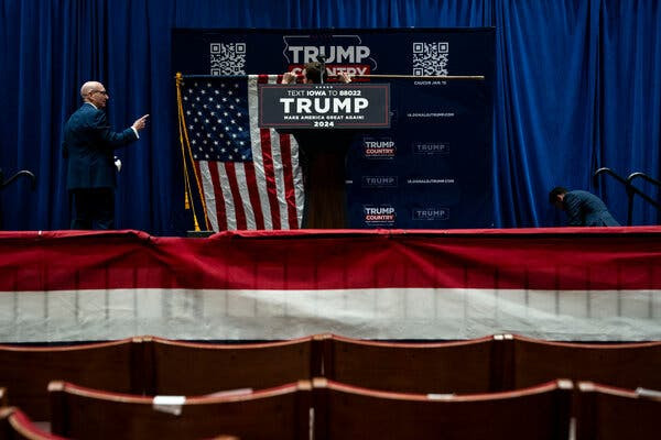 A stage for a Trump rally. There is red and white bunting in the foreground, and a lectern on the stage has a Trump campaign sign. An American flag behind the lectern is being put away.