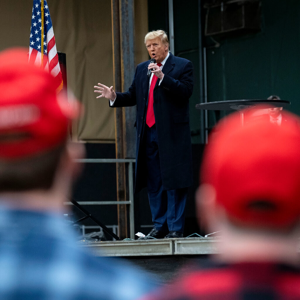 Donald Trump stands on an elevated platform, holding a microphone. People wearing red hats are in the foreground.