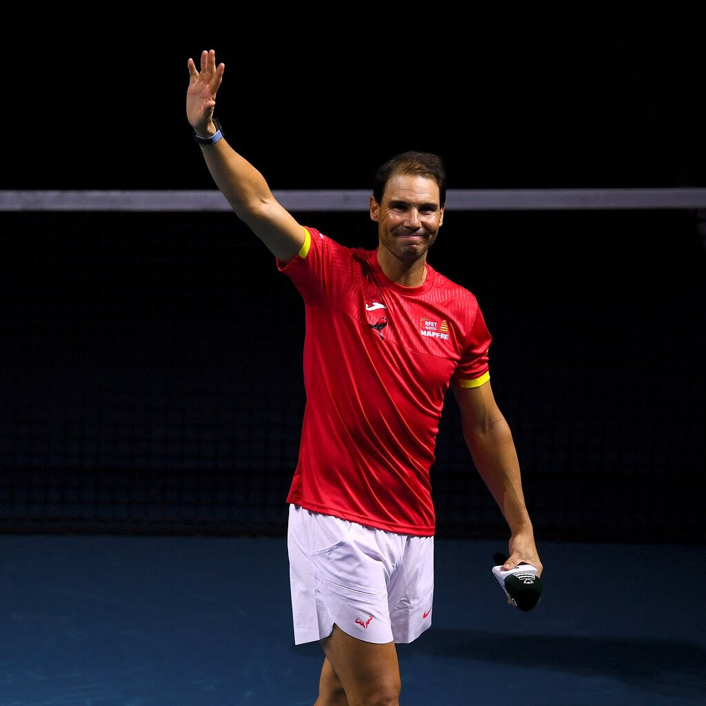 Rafael Nadal in a red top and white shorts waves on court. 